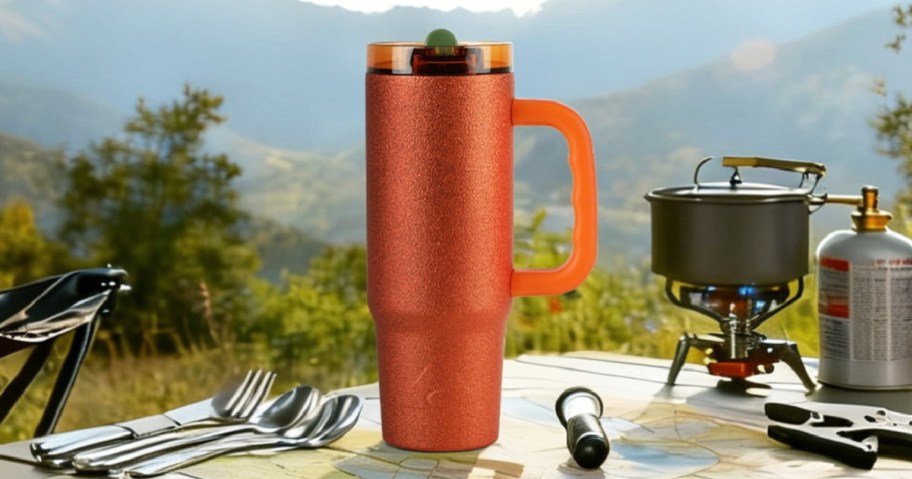 A sparkly orange Ozark Trail 30oz tumbler with a handle resting on a camp table with utensils and a camping stove against a mountain background.