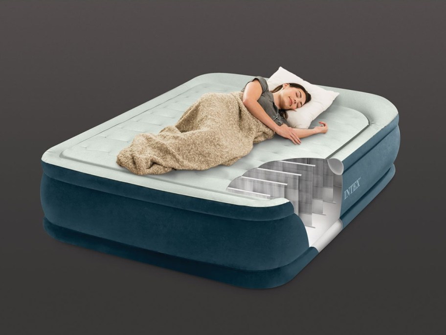 A cutaway diagram of an Intex air mattress showing its internal support structure while a woman sleeps peacefully on top with a blanket and pillow.