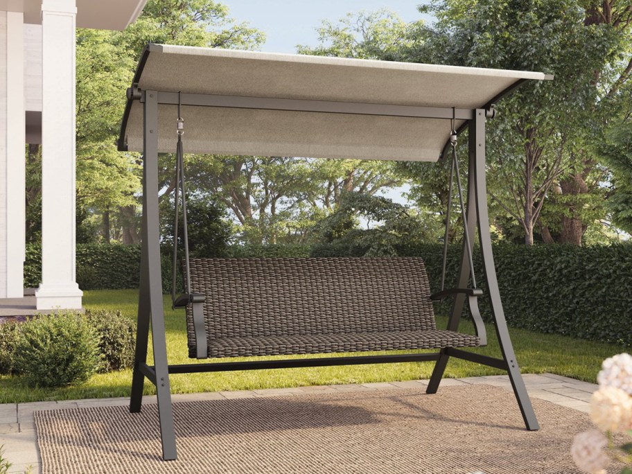 A brown wicker porch swing with a fabric canopy, standing on a patio rug in a backyard.