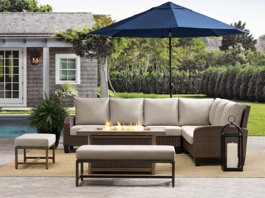 A woven wicker outdoor sectional set with light cushions, a matching fire pit table, benches, and a large blue patio umbrella by a pool.