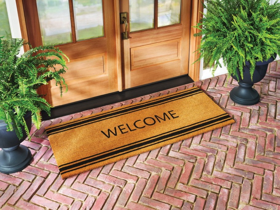 A coir welcome doormat with black stripes on a brick porch in front of double wooden doors, flanked by potted ferns.