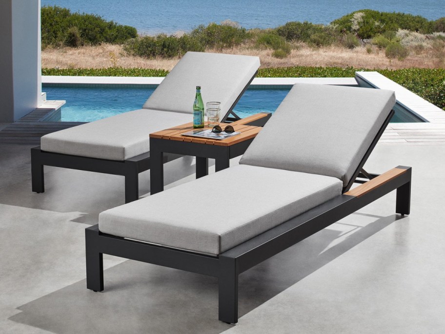 Two black outdoor chaise lounges with light gray cushions and a small side table between them, set by a pool overlooking the water.
