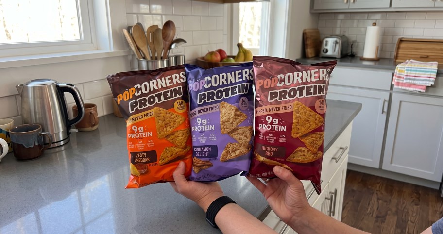 Person holding up 3 PopCorners Protein Chips bags in a kitchen