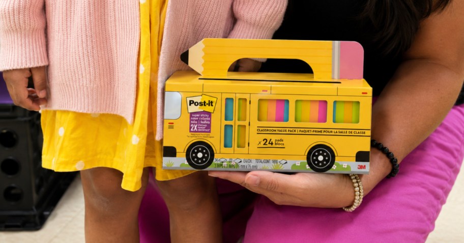 Teacher holding post school bus box and standing next to little girl wearing a dress