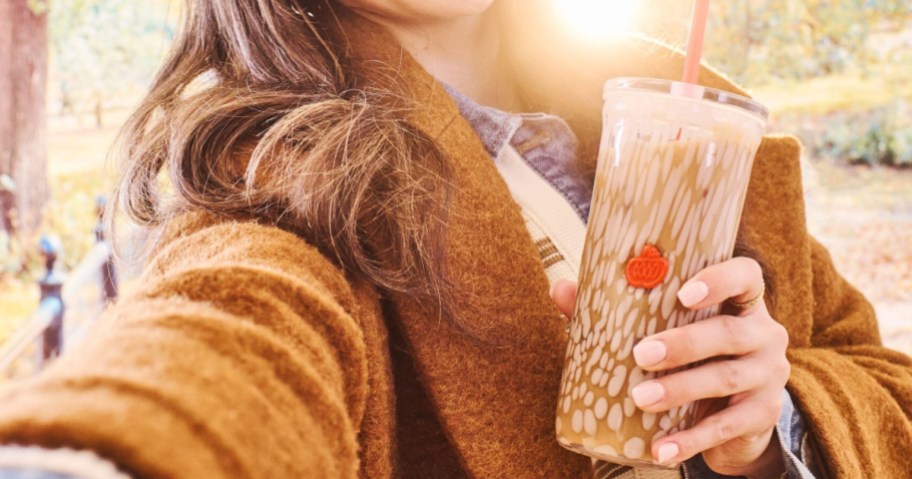 A person wearing a brown coat holds a glass iced coffee cold cup with a decorative pattern and an orange straw outdoors.