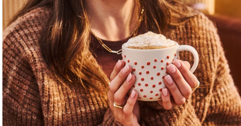 A person wearing a cozy brown sweater holds a white mug with red polka dots filled with a creamy coffee drink topped with whipped cream.