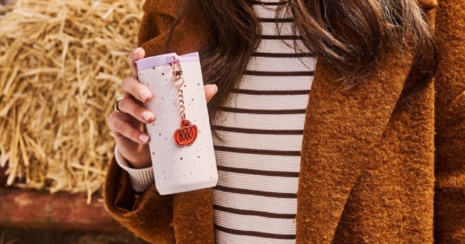 A person wearing a striped shirt and a brown coat holds a light-colored tumbler with colorful speckles and a pumpkin charm attached near hay bales.