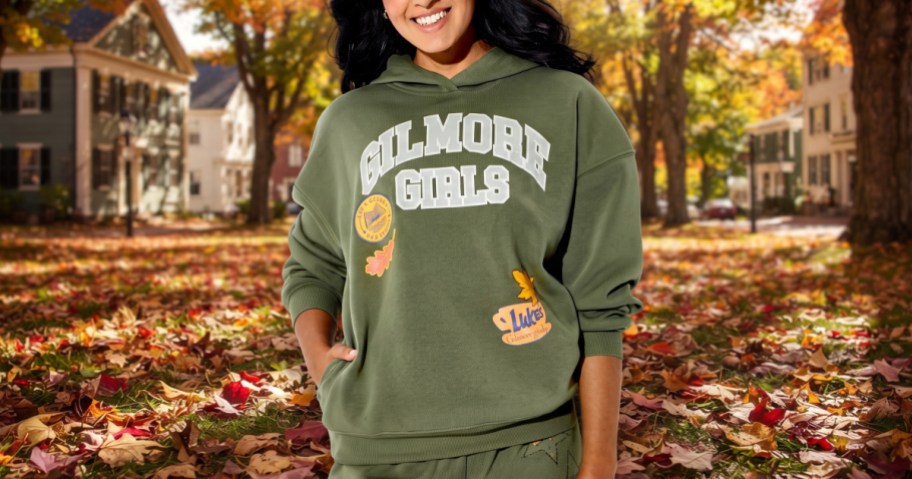 A person wearing an olive green hooded Gilmore Girls patch graphic sweatshirt outdoors against an autumn leaf-strewn background.