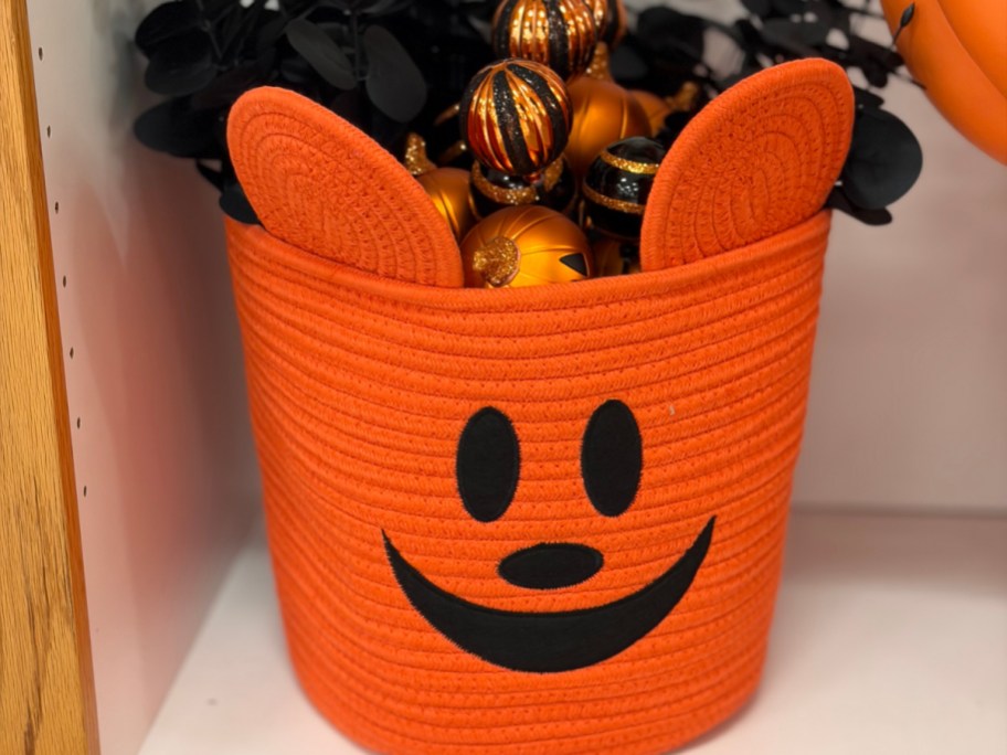 An orange braided storage basket styled like Mickey Mouse's pumpkin face, complete with mouse ears and filled with decorative ornaments.