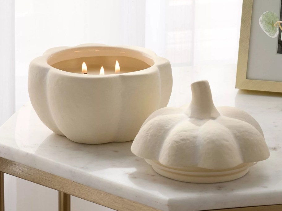 A lit three-wick ceramic pumpkin-shaped candle with its matching lid resting on a marble-top table.