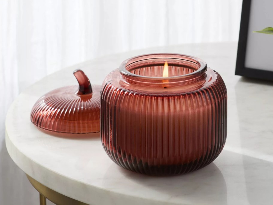 A lit ribbed red glass pumpkin-shaped candle with its matching stem lid resting on a white table.