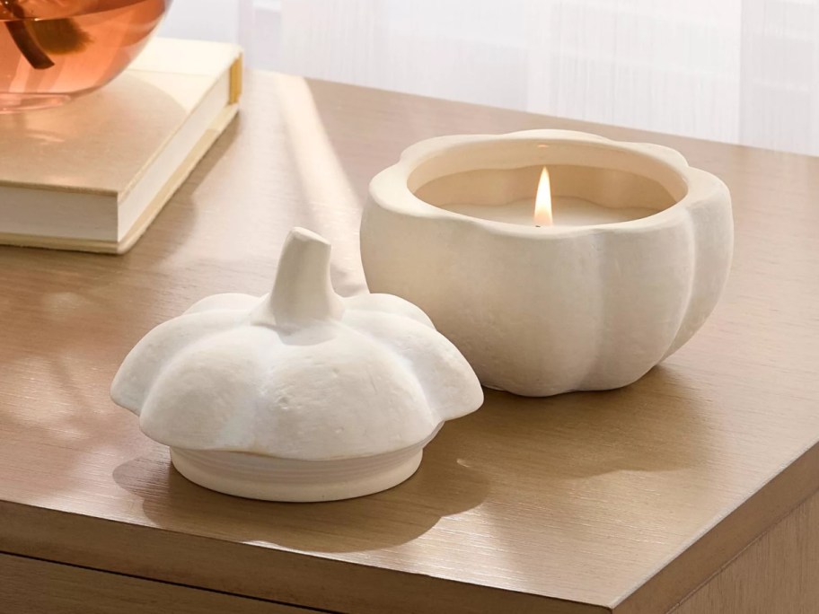 A lit ceramic pumpkin-shaped candle with its matching lid resting on a wooden surface next to a book.