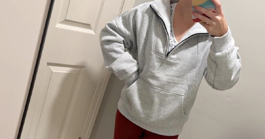 A person wearing an oversized light gray quarter-zip sweatshirt with a front pocket and dark red pants while taking a mirror selfie.