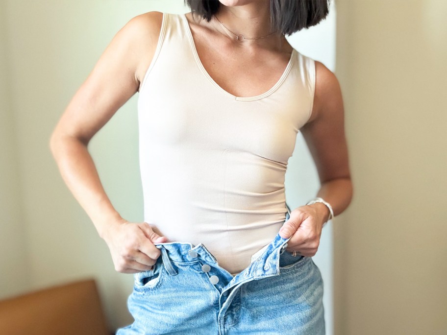 woman pulling on jeans wearing a tan tank top