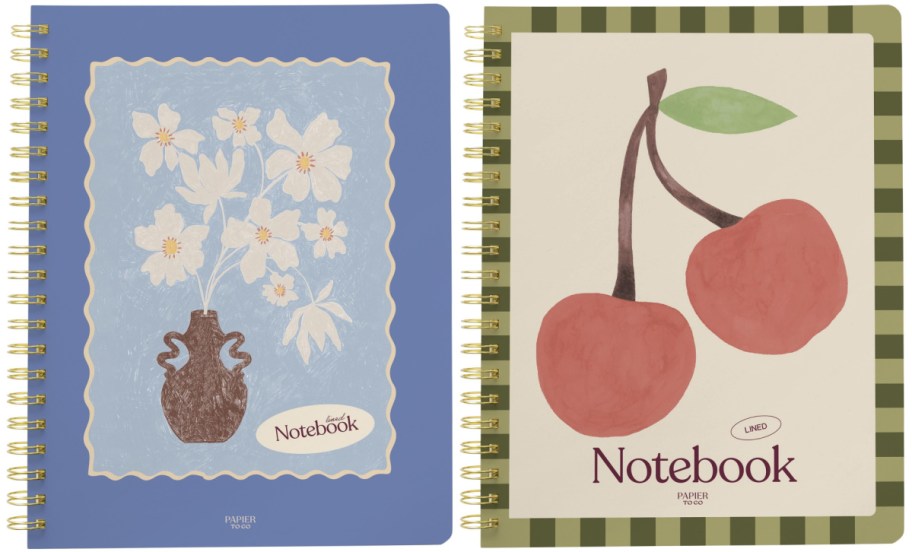 Two Papier to Go spiral-bound lined notebooks featuring a blue cover with a white floral vase painting on the left and a cream cover with a green-trimmed checkered border and a large cherry illustration on the right.
