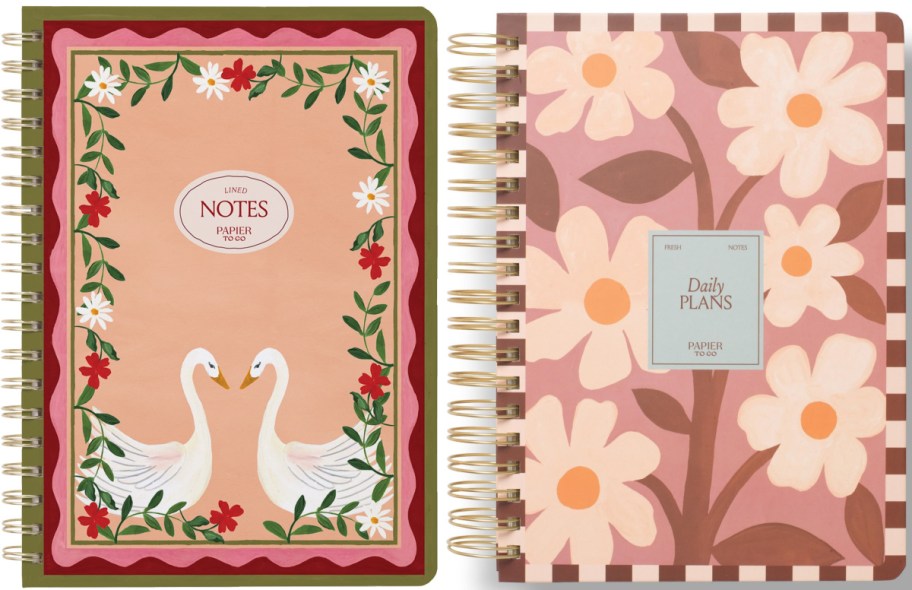 Two Papier to Go spiral-bound notebooks featuring a peach cover with a floral border and two swans facing each other on the left, and a pink cover with large white and orange flowers and a checkered border on the right.