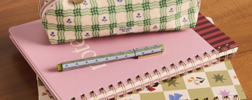 Papier to Go stationery items including a green plaid pencil pouch, a patterned pen, and spiral-bound notebooks stacked on a wooden surface.