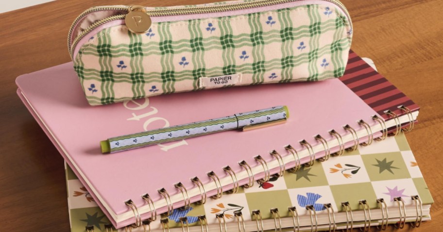 Papier to Go stationery items including a green plaid pencil pouch, a patterned pen, and spiral-bound notebooks stacked on a wooden surface.