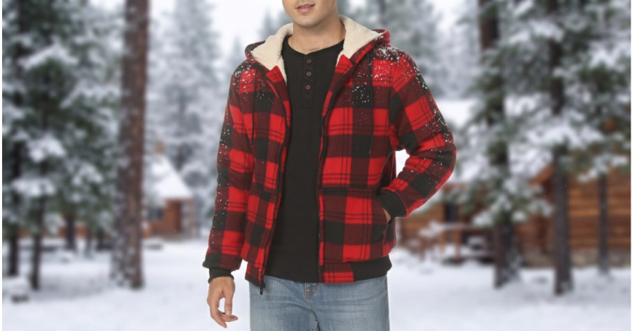 A person wearing a red and black plaid sherpa-lined hooded jacket over a black shirt and jeans in a snowy outdoor setting.