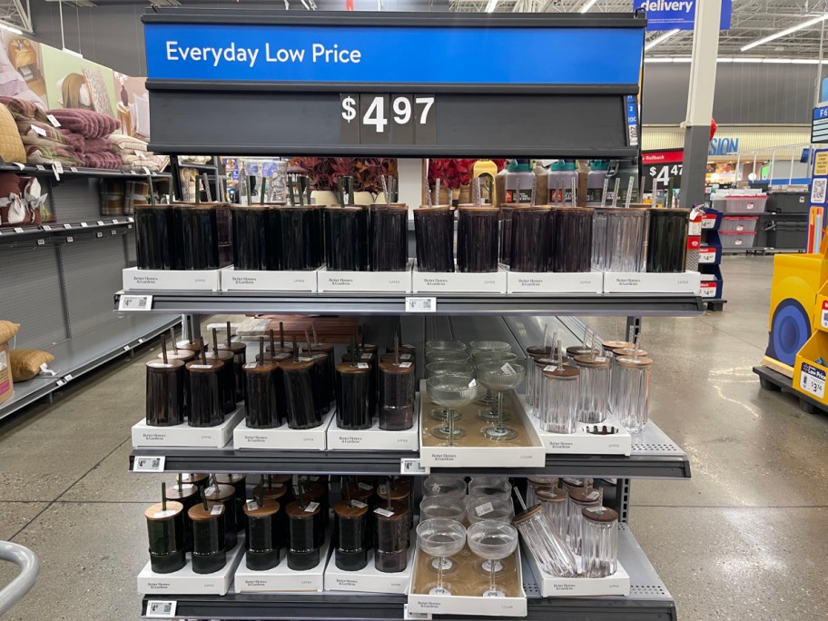 A retail store display featuring multiple shelves of Better Homes & Gardens ribbed glass tumblers with wooden lids and straws, alongside clear ribbed coupe glasses, beneath a blue Everyday Low Price sign for $4.97.