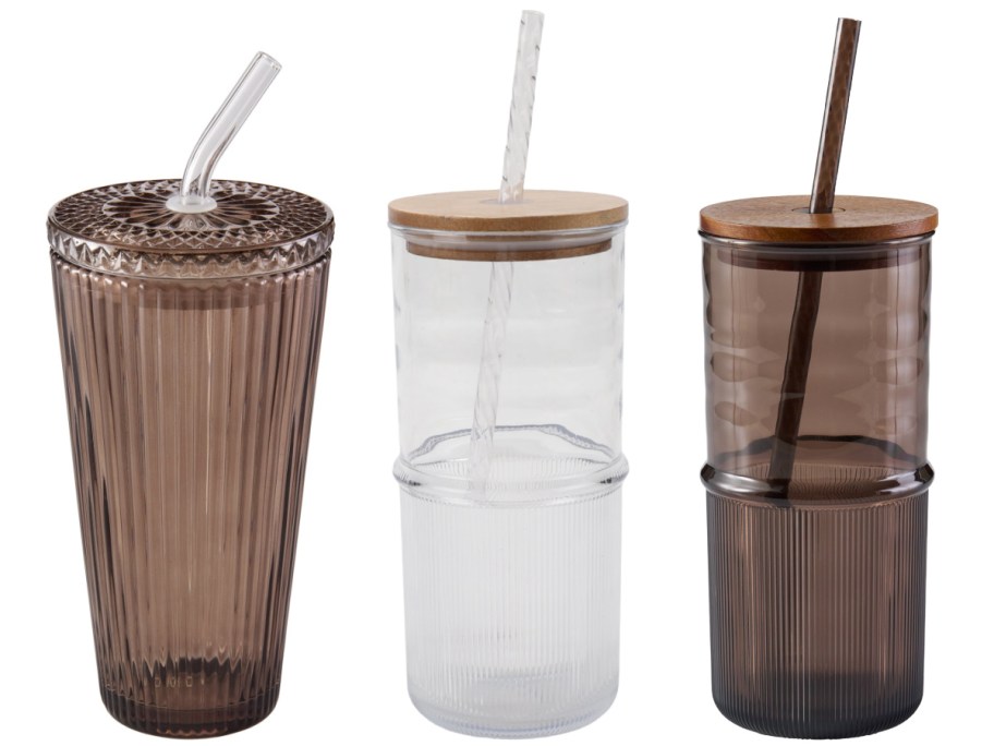 Three glass tumblers with lids and straws side-by-side on a white background. The left tumbler is entirely ribbed amber glass with a matching lid, while the middle clear tumbler and right amber tumbler both have wooden lids and ribbed bottom halves.
