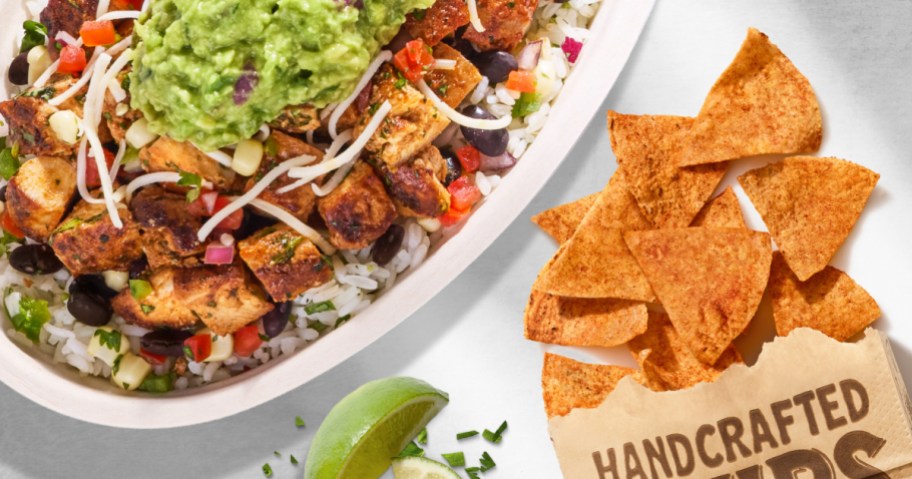 A Chipotle bowl with chicken, rice, beans, and guacamole next to a bag of handcrafted chips and lime wedges.