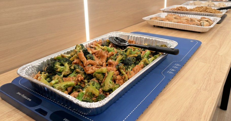FJAU blue food warming mat on a wooden table holding a pan of cooked broccoli and chicken.