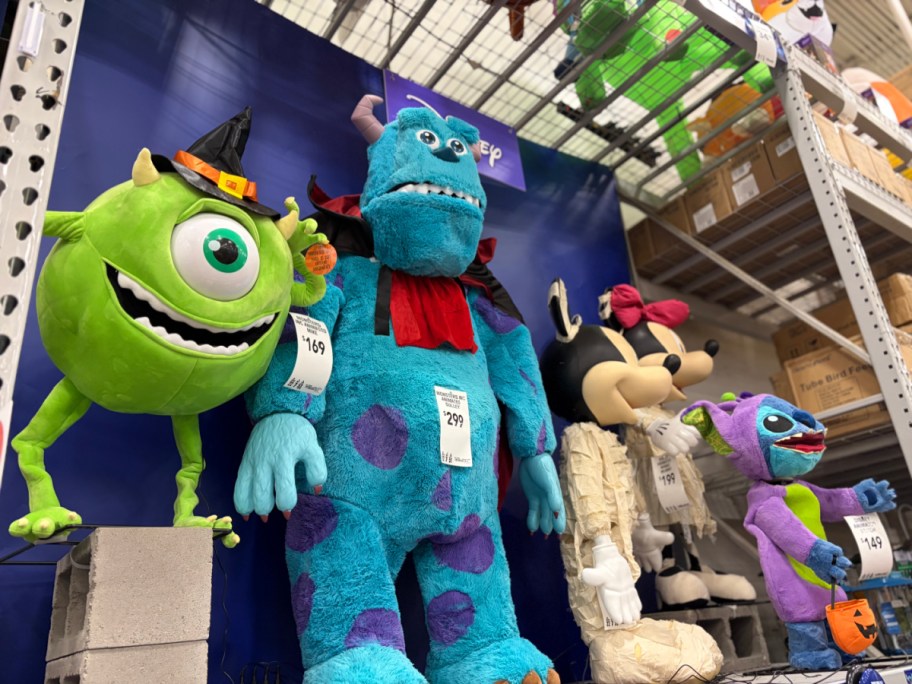 A display of Halloween animatronics at a store, featuring Mike Wazowski wearing a witch hat on the left, a large Sulley animatronic in a cape, a mummy character, and Stitch holding a pumpkin bucket on the right.