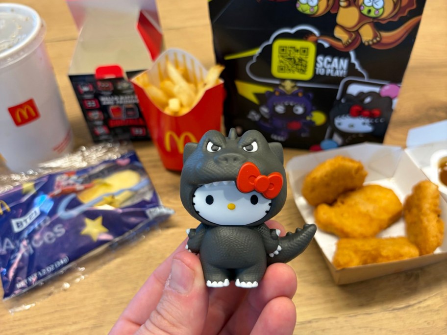 hand holding a hello kitty dressed as godzilla in front of the contents of a happy meal