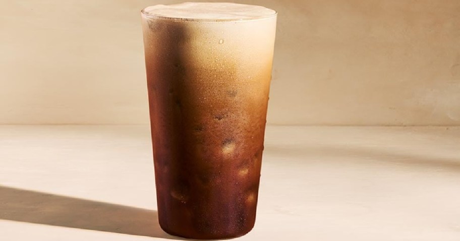 An iced Starbucks Americano in a tall clear glass with a layer of foam on top.