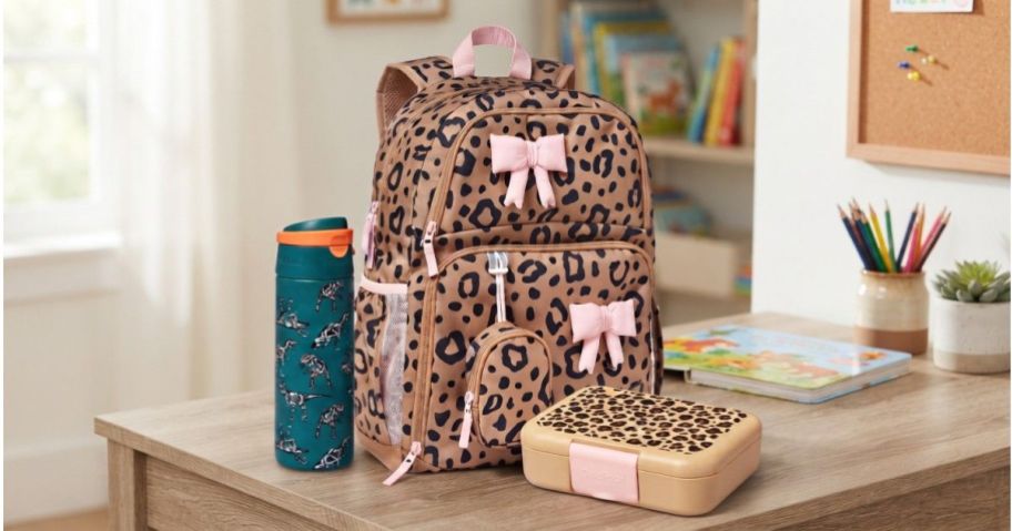 A cheetah-print backpack decorated with pink bows, a matching lunch box, and a teal water bottle with dinosaur prints, sitting on a wooden desk.