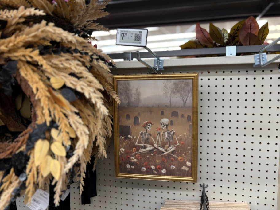 A framed painting of two skeletons sitting in a graveyard, hanging next to a decorative fall wreath with black bats.