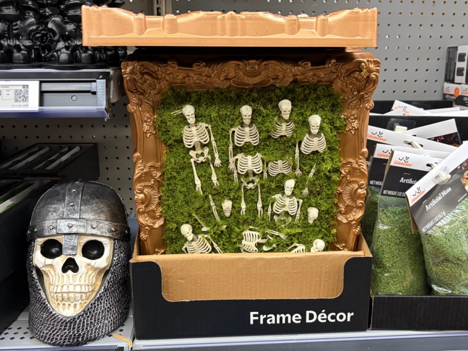 An ornate gold-framed Halloween decor piece filled with green moss and multiple miniature skeleton figures, displayed on a store shelf next to a skull wearing a medieval helmet.