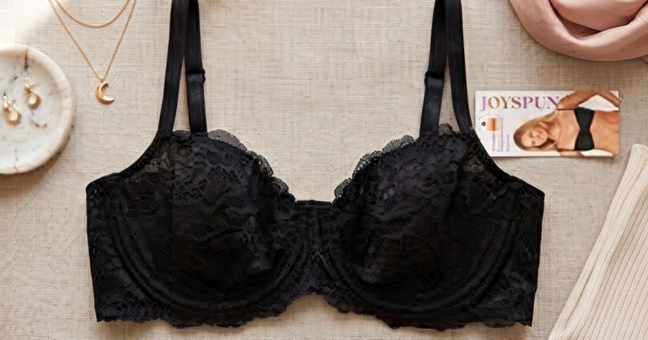 A black lace bra displayed flat on a textured surface alongside jewelry, a brand tag, and clothing pieces.