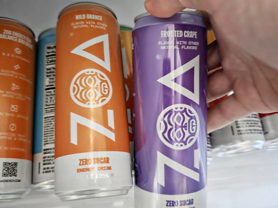A hand holding a purple Frosted Grape ZOA energy drink can next to an orange Wild Orange can in a refrigerator.