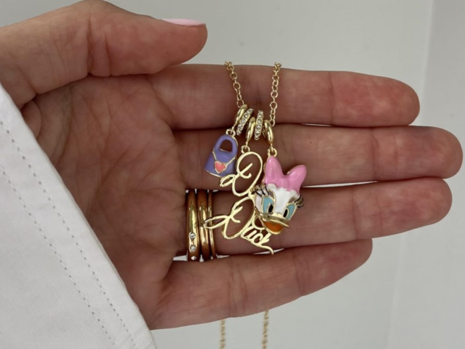 A gold Disney character charm necklace featuring Daisy Duck, a purple handbag charm, and script lettering charms displayed on a person's hand.