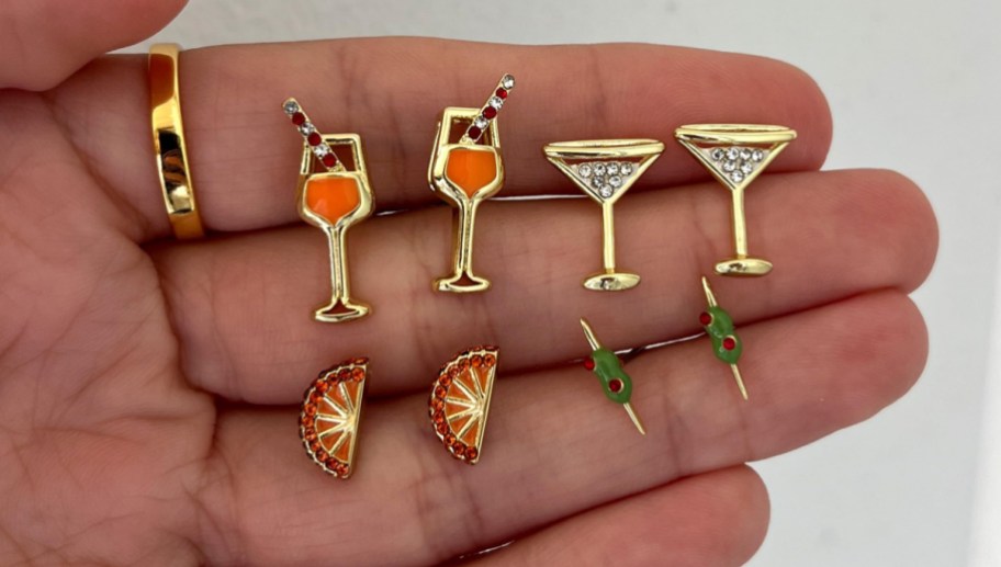 A set of gold novelty cocktail-themed stud earrings featuring miniature drink glasses with straws, martini glasses filled with rhinestones, olive picks, and orange wedge slices displayed on a person's hand.