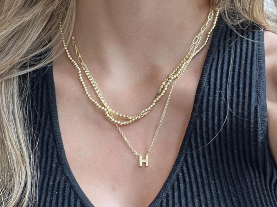 A person wearing layered gold chain necklaces, including a delicate chain with a letter "H" initial pendant.