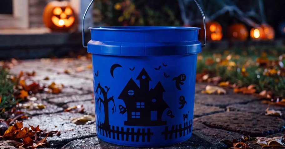 A blue plastic Halloween bucket with a black spooky haunted house silhouette and wire handle, sitting on a pathway with autumn leaves.