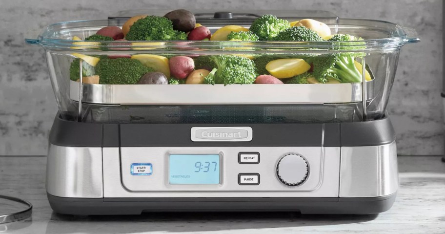 A Cuisinart super food steamer filled with fresh vegetables like broccoli, potatoes, and squash in a clear glass container on top of a digital base.