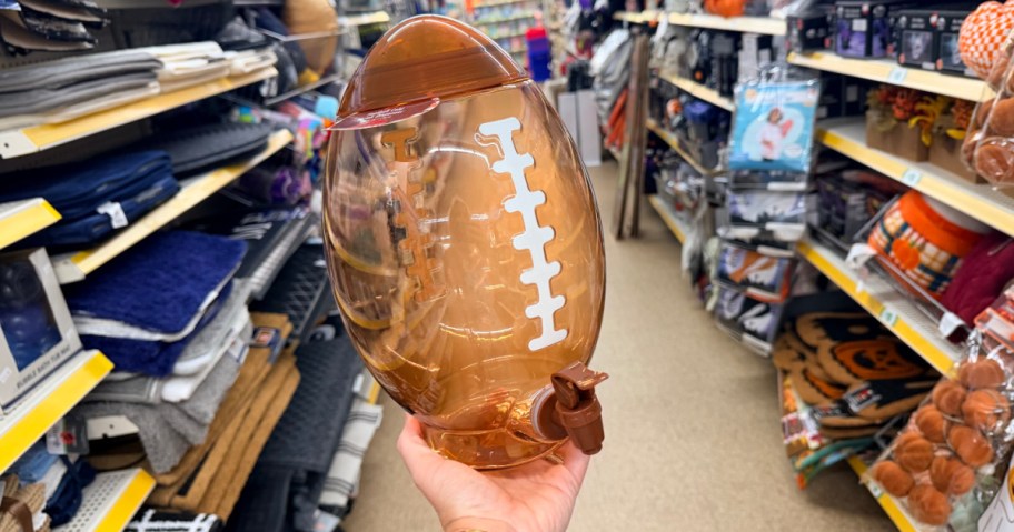A brown football-shaped drink dispenser with a spigot and white laces, held up in a store aisle.