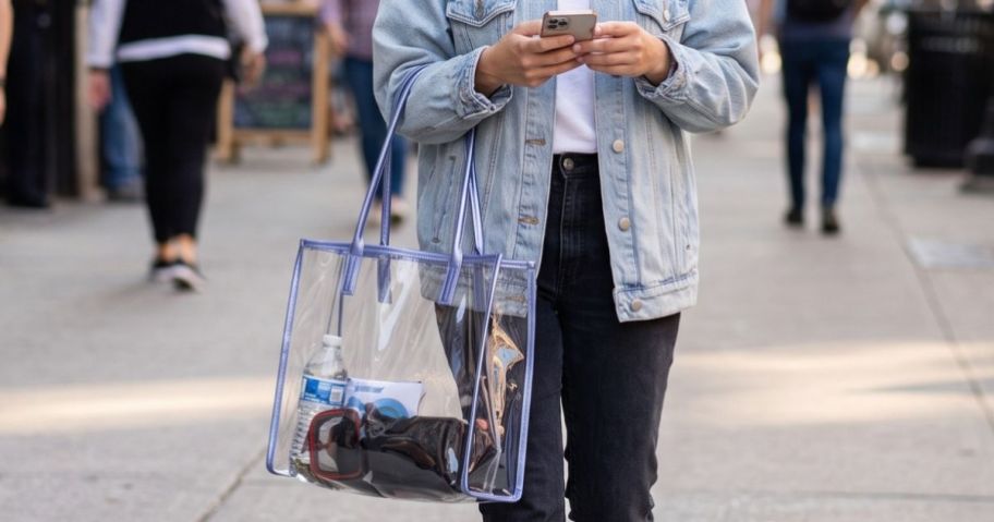 Woman carrying a clear shopper tote bag filled with essetials while walking down a sidewalkk on her phone