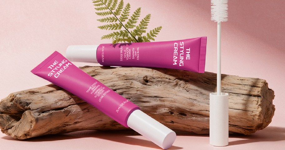 Two pink tubes of styling cream displayed on a wooden log with a mascara-style application brush.