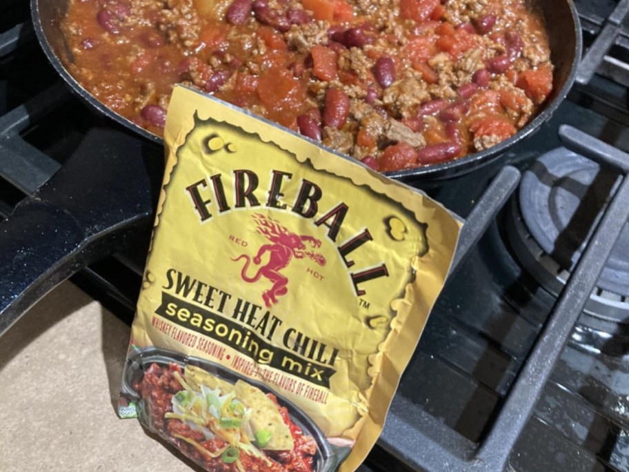 A packet of Fireball Sweet Heat Chili seasoning mix placed next to a skillet filled with cooking chili on a stovetop.