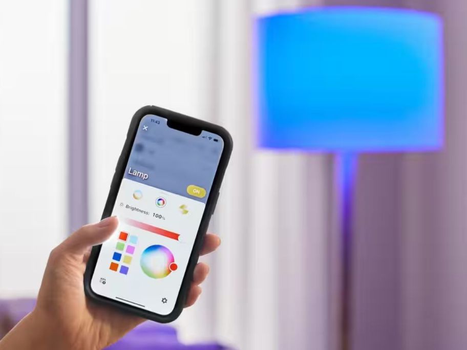 Hand holding a phone with an app to control a smart bulb on the screen with a lamp with a blue colored bulb in the background