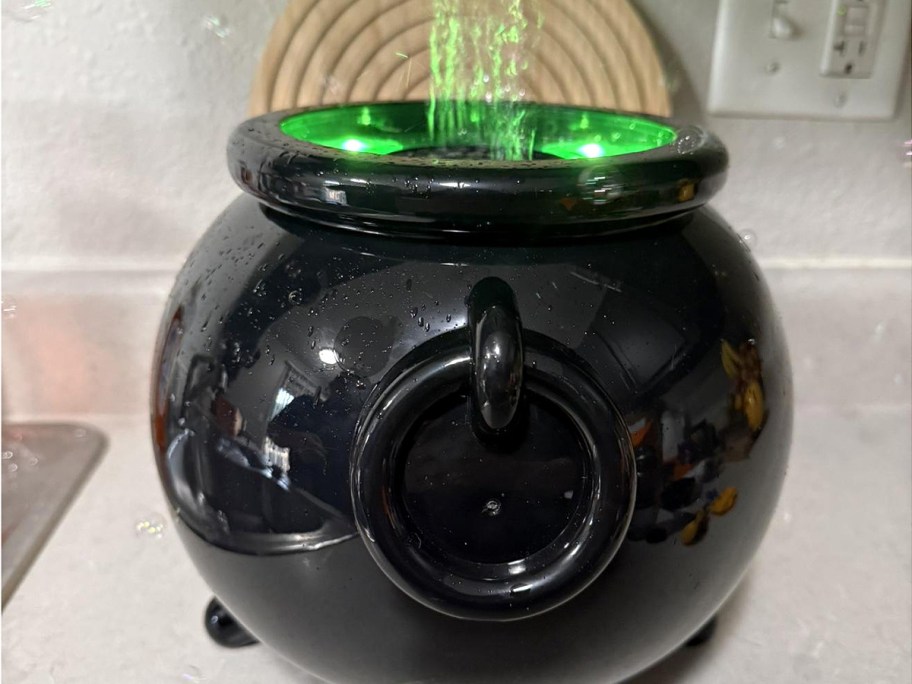 A black plastic cauldron bubble blower with green light glowing from inside and bubbles blowing out the top on a countertop.