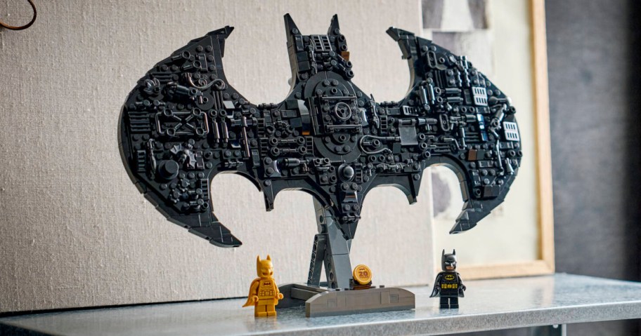 A black LEGO DC Batman Logo built from bricks on a stand, accompanied by two Batman minifigures standing at the base on a shelf.