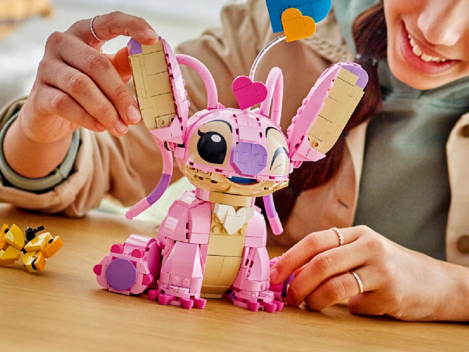 A person building a pink and tan LEGO Disney Angel figure from Lilo and Stitch on a wooden table.
