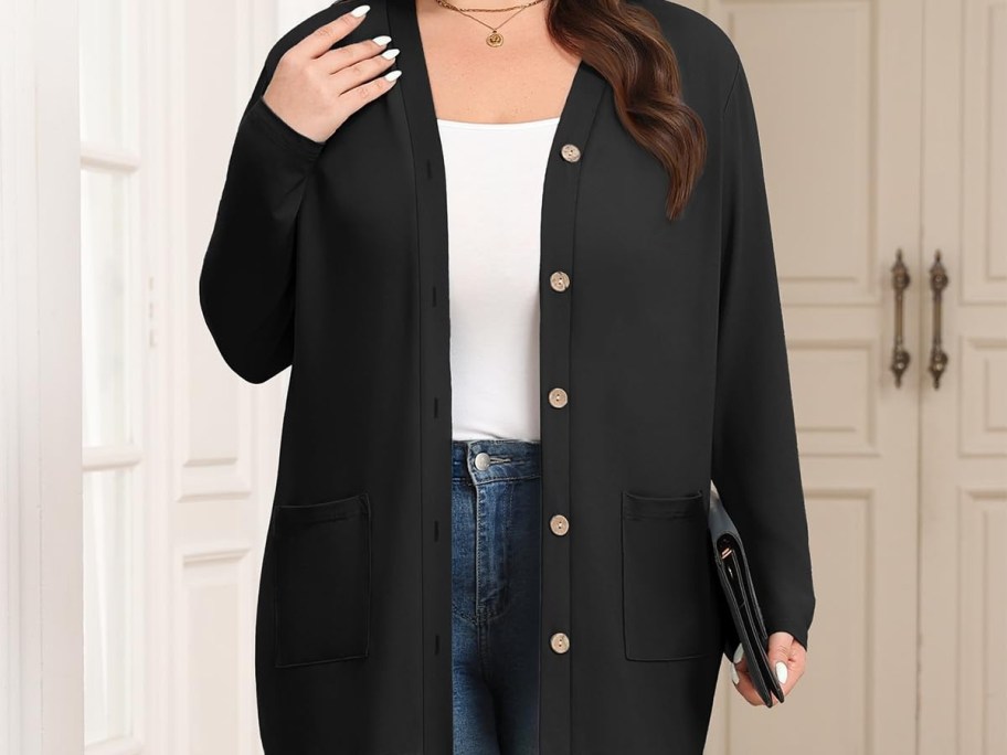 A woman wearing an open black button-down cardigan with pockets over a white top and blue jeans, holding a small clutch.