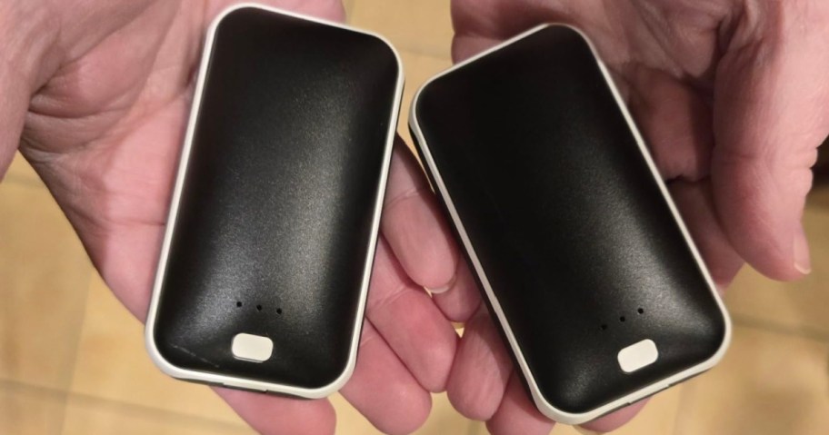 A person holding two black and white rechargeable hand warmers with indicator lights and power buttons.